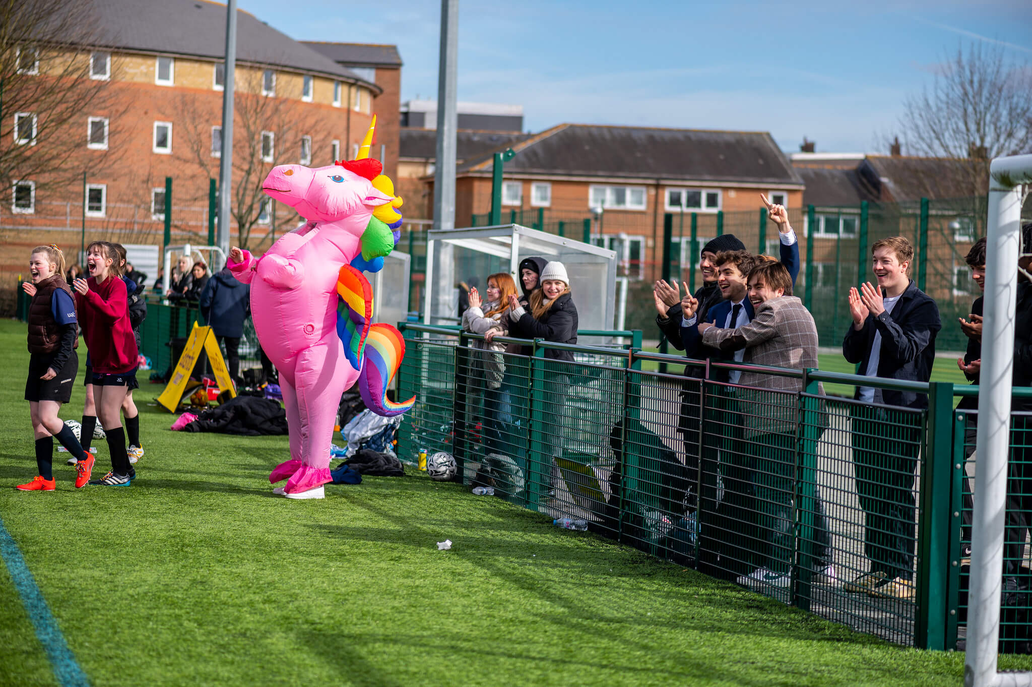 A pink unicorn stands clapping on a field of grass with spectators behind a fence.
