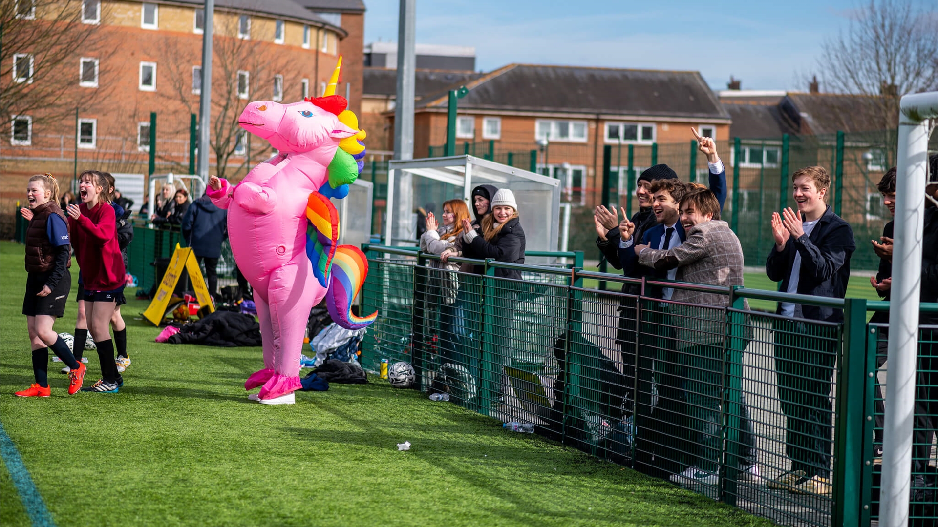 A pink unicorn stands clapping on a field of grass with spectators behind a fence.