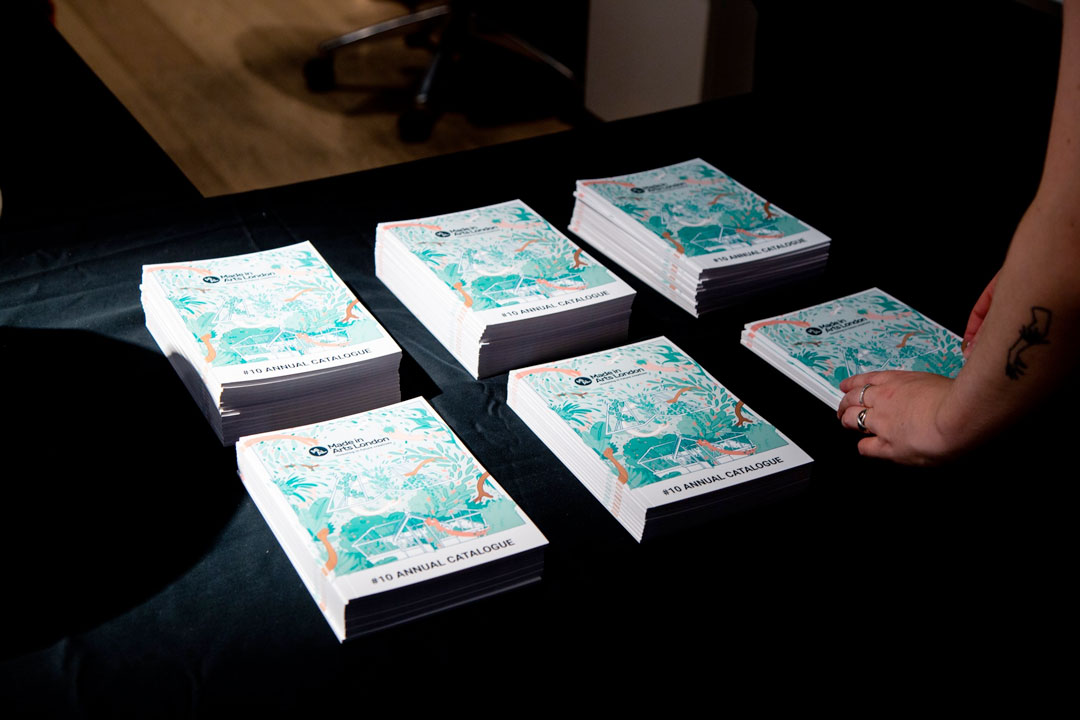 A photograph of copies of the Made in Arts London Annual Catalogue laid out on a table with a person looking at them