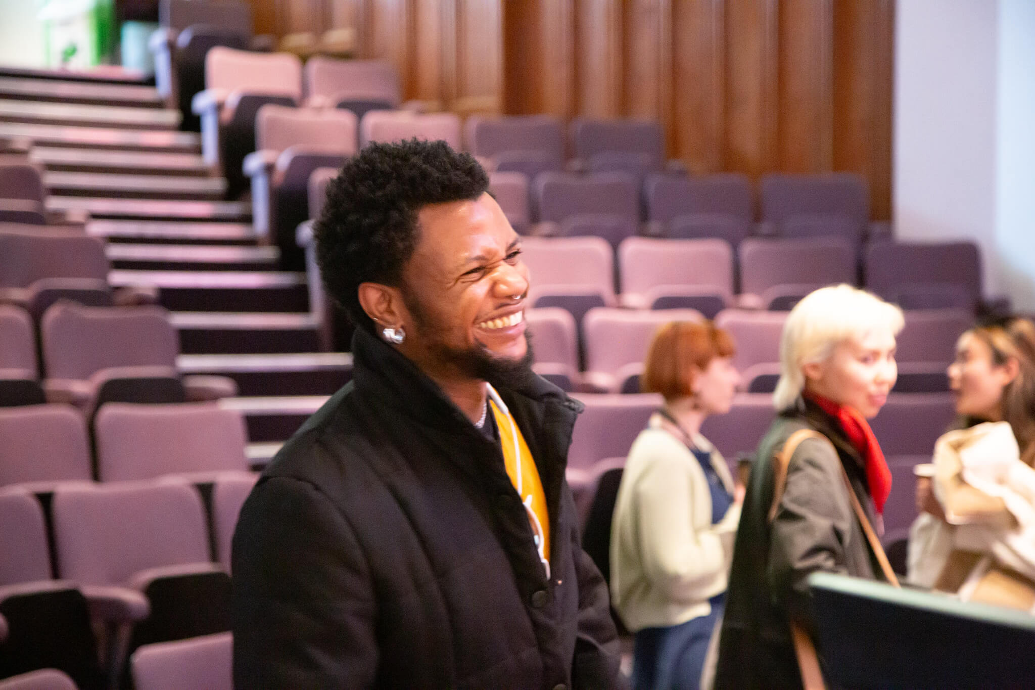 A person smiling, they are wearing a black coat with a yellow shirt underneath.