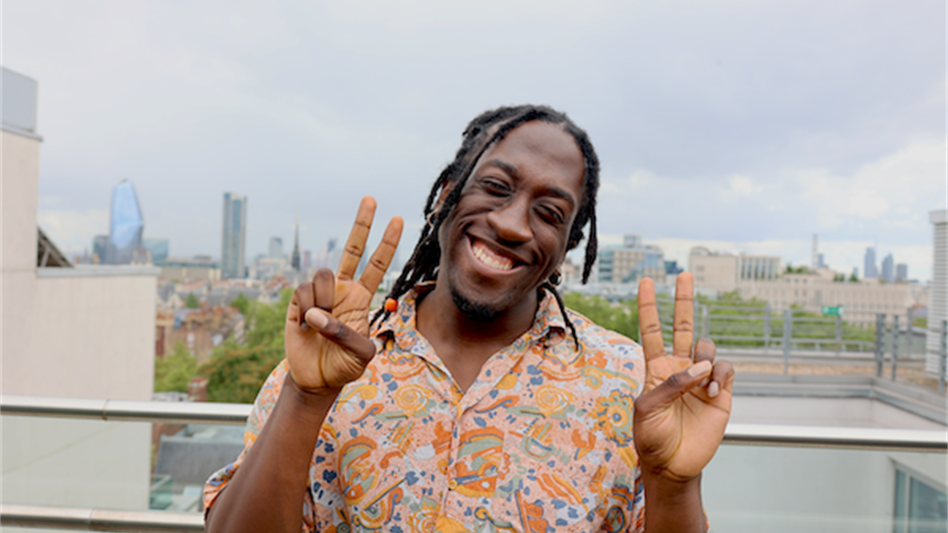 A person with dark brown dreadlocks, smiling holding up two "peace" signs with their hands. They're wearing a multicoloured short sleeved shirt.