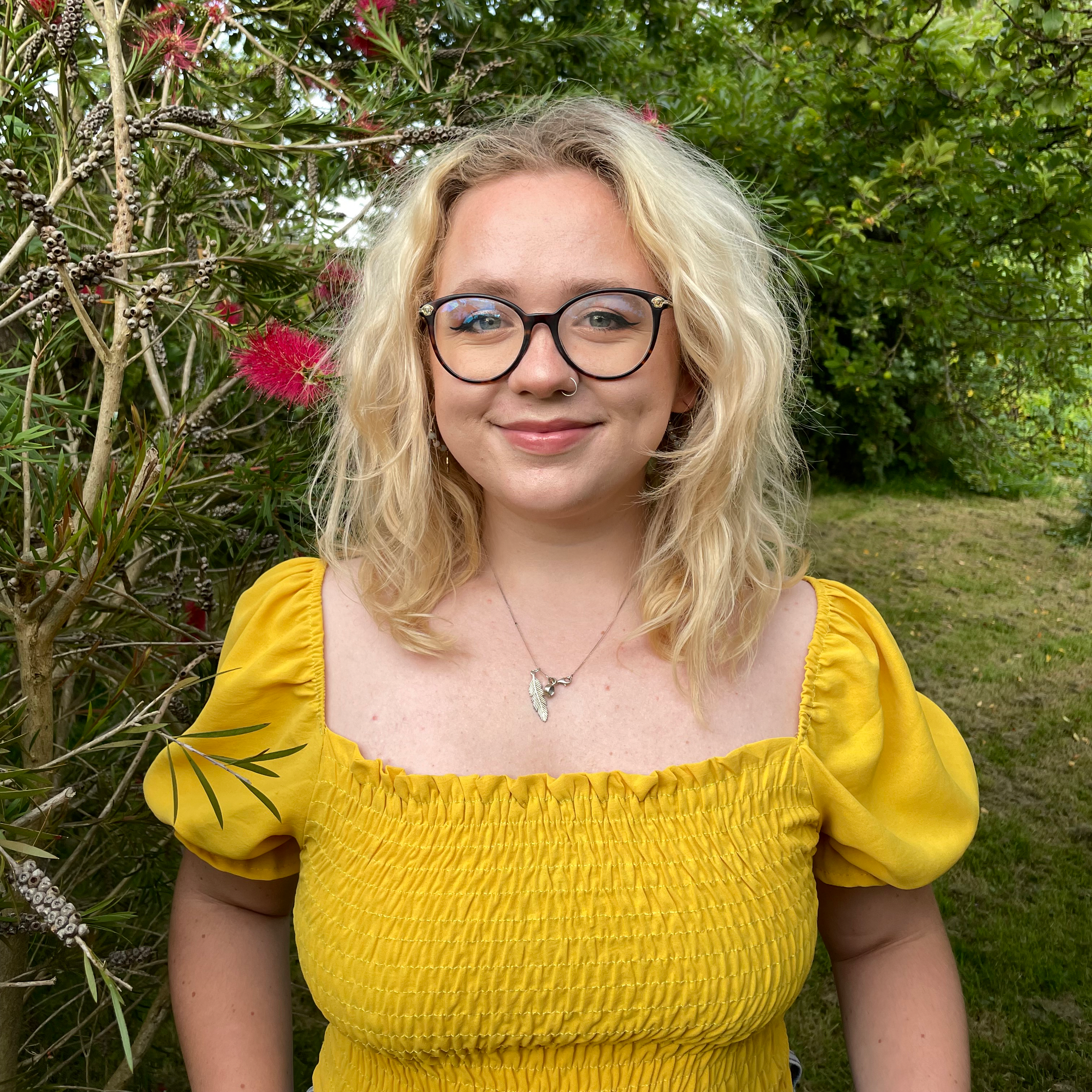 A photo of Georgia, who is wearing black glasses, a necklace, and a yellow top.